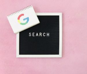 pink background with black and white letter board that says search in white letters and google logo on spiral notebook in left hand corner