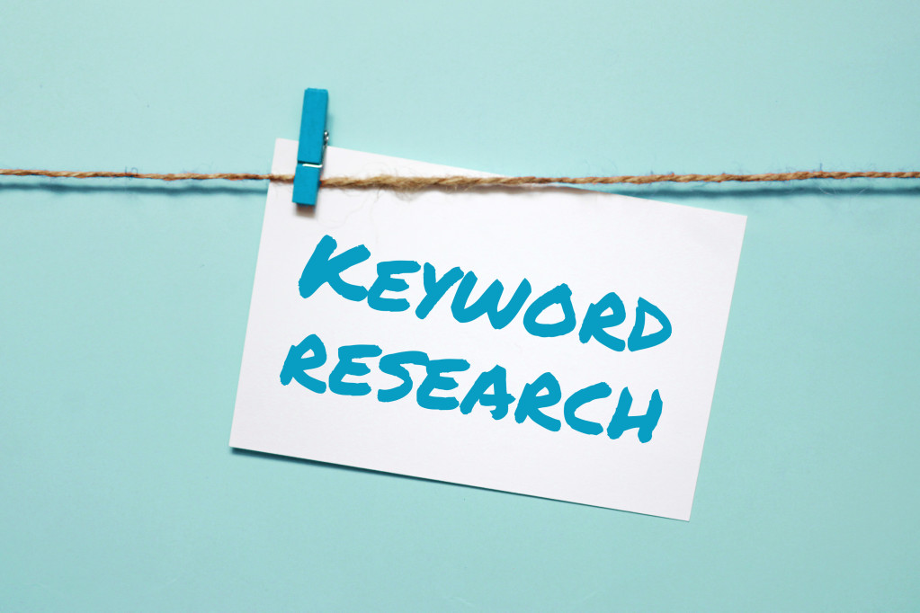Keyword research written in blue on white paper being held up by blue close-in on close line with blue background