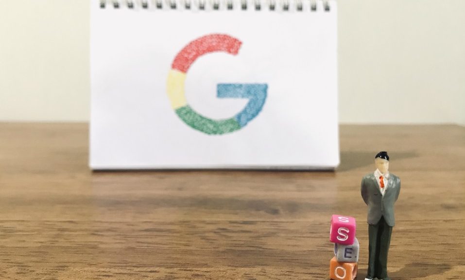 Google logo on spiral notebook in background on wooden table and little figurine with three bead letters next to him that spell SEO